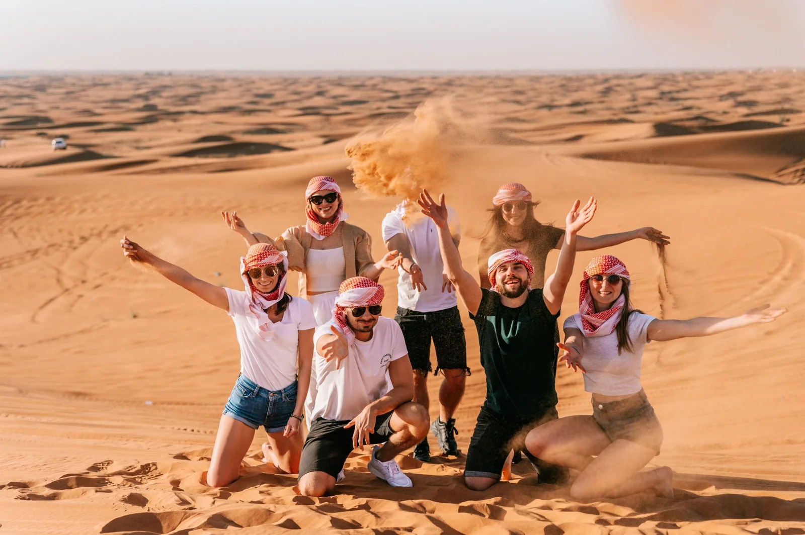 Group of friends wearing headscarves and sunglasses joyfully tossing sand in the air while kneeling on golden desert dunes under a clear sky.